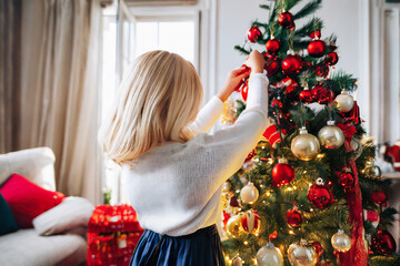 Little blonde haired girl decorating big Christmas tree with red and gold decorations in living room. Captured from behind with selective focus. ai generated