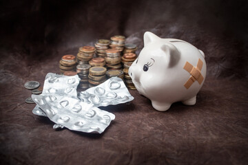 White ceramic piggybank next to pill blisters and stacks of coins on brown leather background