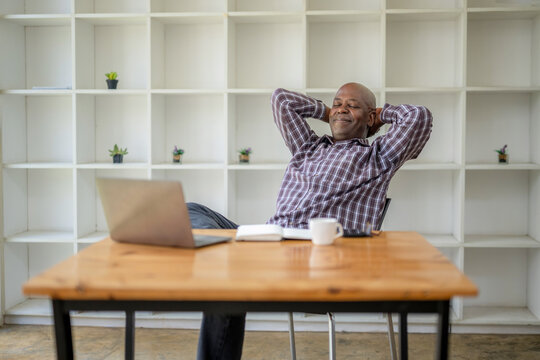 Senior black man puts his hands behind his head. Leaning on an office chair. Take a break from working in front of laptop screen.