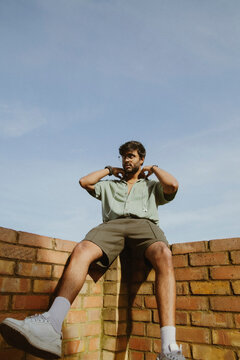 Young Indian guy in trendy clothes sitting on brick wall with earphones around neck