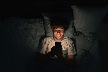 Young sleepy exhausted man lying in bed using smartphone at late night. Insomnia, sleep disorder concept. Addiction on a cell phone. - obrazy, fototapety, plakaty