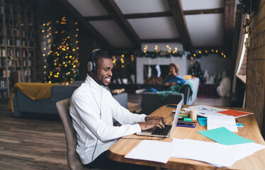 Black male in headphones sits at wooden desk, typing on a laptop in cozy, festive home office