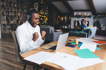 Obraz premium Excited Black man in a white turtleneck with headphones at a wooden desk celebrates success on his laptop