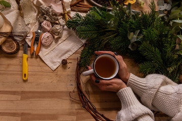 Top view still life, flat lay female hands holding white cup of tea on wooden table. Place for text, on background are tools, natural materials for making handmade Christmas wreath. Copy space