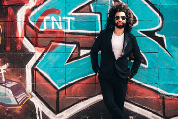 Handsome hipster model. Arabian man dressed in suit jacket clothes. Fashion male with long curly hairstyle posing in street near graffiti wall