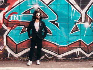 Handsome hipster model. Arabian man dressed in suit jacket clothes. Fashion male with long curly hairstyle posing in street near graffiti wall