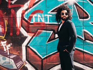 Handsome hipster model. Arabian man dressed in suit jacket clothes. Fashion male with long curly hairstyle posing in street near graffiti wall