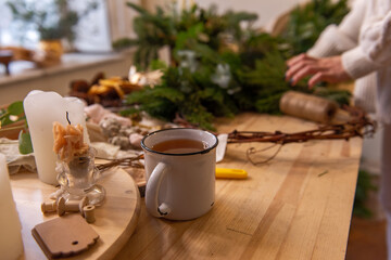 Side view still life, flat lay female hands holding white cup of tea on wooden table. Place for text, on background are tools, natural materials for making handmade Christmas wreath. Copy space