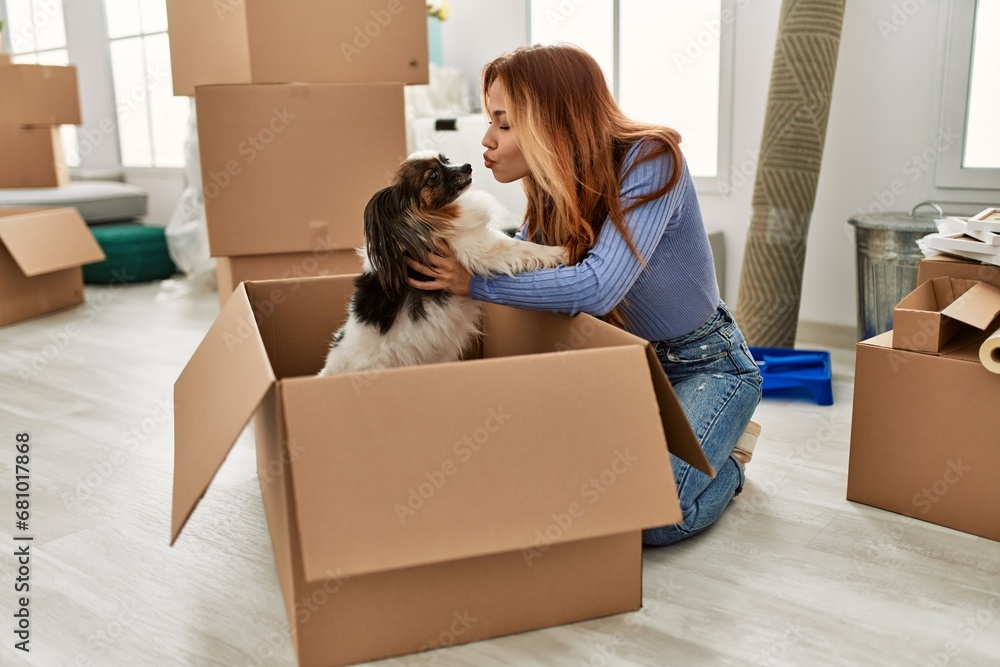 Sticker Young caucasian woman sitting on floor holding dog of cardboard box at new home