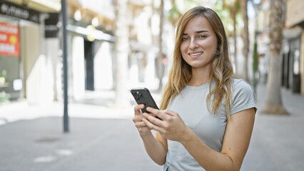 Cheerful young blonde woman happily touching her smartphone, engaged in a digital conversation on a sunny city street