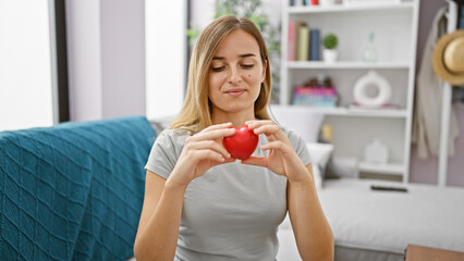 Confident young adult woman, enjoy the indoors lifestyle, sitting comfortably on her cozy living room sofa, holding a heart, with a big smile lighting up her beautiful, blonde portrait.