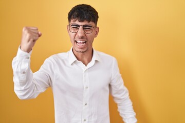 Young hispanic man standing over yellow background angry and mad raising fist frustrated and...