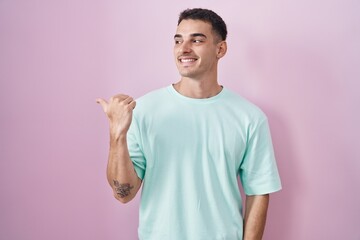Handsome hispanic man standing over pink background smiling with happy face looking and pointing to the side with thumb up.