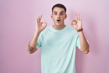 Handsome hispanic man standing over pink background looking surprised and shocked doing ok approval symbol with fingers. crazy expression