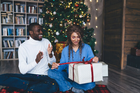 cheerful interracial couple in casual attire unwraps a Christmas present