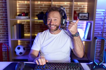 Middle age man with beard playing video games wearing headphones doing happy thumbs up gesture with...