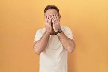 Middle age man with beard standing over yellow background rubbing eyes for fatigue and headache,...