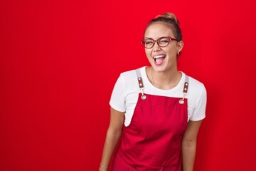 Young hispanic woman wearing waitress apron over red background winking looking at the camera with...