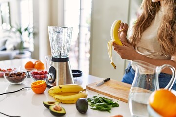 Young beautiful hispanic woman preparing vegetable smoothie with blender peeling banana at the kitchen - obrazy, fototapety, plakaty