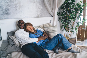 Relaxed and happy interracial couple cuddling on a bed with comfortable cushions, in a cozy
