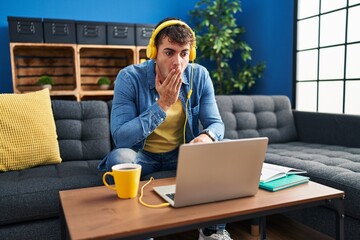 Young hispanic man using laptop at home covering mouth with hand, shocked and afraid for mistake....