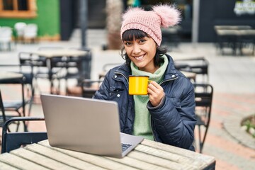 Young beautiful hispanic woman using laptop drinking coffee sitting on table at coffee shop terrace