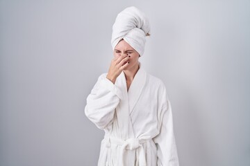 Blonde caucasian woman wearing bathrobe smelling something stinky and disgusting, intolerable smell, holding breath with fingers on nose. bad smell