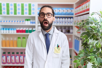 Hispanic man with beard working at pharmacy drugstore scared and amazed with open mouth for surprise, disbelief face