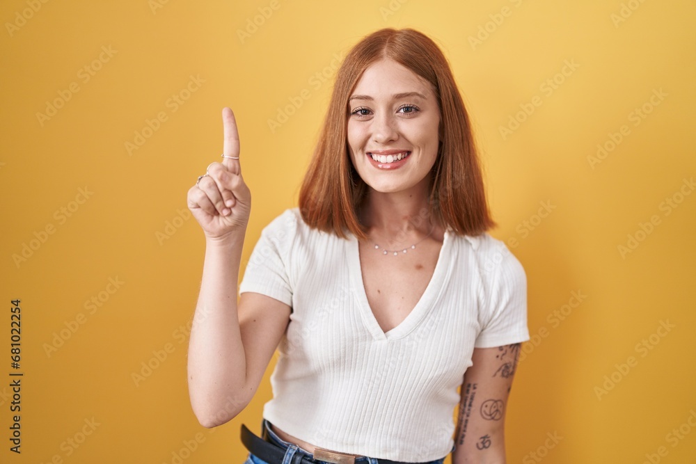 Sticker Young redhead woman standing over yellow background showing and pointing up with finger number one while smiling confident and happy.