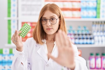 Young redhead woman working at pharmacy drugstore holding birth control pills with open hand doing stop sign with serious and confident expression, defense gesture - obrazy, fototapety, plakaty