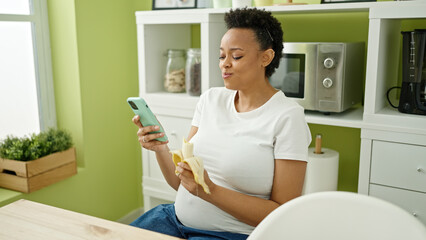 Young pregnant woman eating banana using smartphone at dinning room