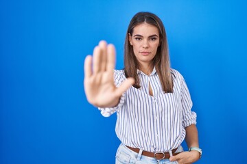 Hispanic young woman standing over blue background doing stop sing with palm of the hand. warning expression with negative and serious gesture on the face.