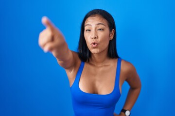 Fototapeta na wymiar Hispanic woman standing over blue background pointing with finger surprised ahead, open mouth amazed expression, something on the front
