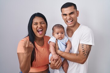Young hispanic couple with baby standing together over isolated background angry and mad raising...