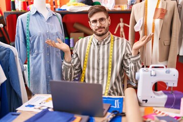 Hispanic man with beard using laptop at tailor room clueless and confused expression with arms and hands raised. doubt concept.