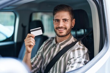 Hispanic man with beard driving car holding credit card looking positive and happy standing and smiling with a confident smile showing teeth