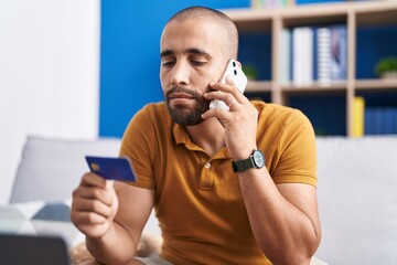 Young latin man talking on smartphone holding credit card with serious expression at home