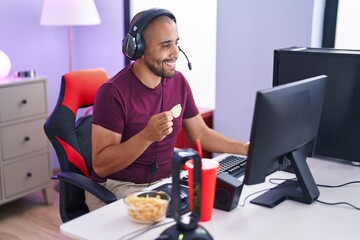 Young latin man streamer playing video game eating chips potatoes at gaming room