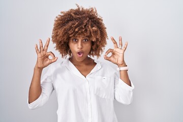 Young hispanic woman with curly hair standing over white background looking surprised and shocked doing ok approval symbol with fingers. crazy expression