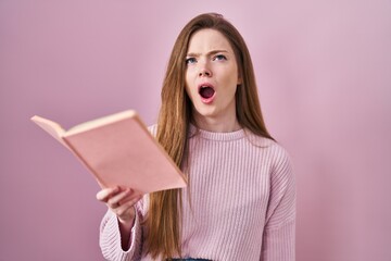 Young caucasian woman reading a book over pink background angry and mad screaming frustrated and furious, shouting with anger looking up.