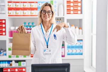 Young blonde woman working at pharmacy drugstore holding paper bag pointing finger to one self...