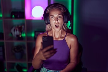Young blonde woman playing video games with smartphone scared and amazed with open mouth for surprise, disbelief face