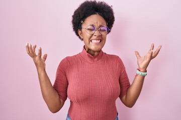 Beautiful african woman with curly hair standing over pink background celebrating mad and crazy for...