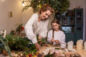 Young mother and little daughter together make Christmas wreath from spruce, eucalyptus branches....