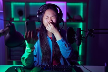 Young asian woman playing video games with smartphone smelling something stinky and disgusting,...