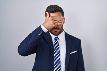 Handsome hispanic man wearing suit and tie covering eyes with hand, looking serious and sad. sightless, hiding and rejection concept