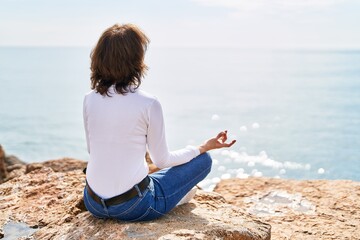 Naklejka na ściany i meble Middle age woman doing yoga exercise sitting on the rock at seaside