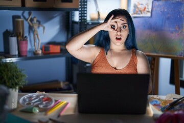 Young modern girl with blue hair sitting at art studio with laptop at night doing ok gesture shocked with surprised face, eye looking through fingers. unbelieving expression.