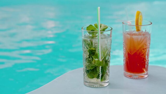 Two cold fresh made cocktails. Mojito and aperol glass on poolside made by barman. Mid shot of alcohol beverage with straws and ice cubes. Swimming pool alco party