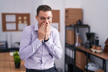 Young hispanic man at the office laughing and embarrassed giggle covering mouth with hands, gossip...